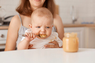 Faceless mother feeds her little infant daughter with vegetable puree, cute child trying to eat by her own, eating tasty healthy food, feeding up, indoor shot.