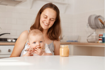 Portrait of smiling happy woman sitting at table in light room with her infant daughter, looking at baby with smile, adorable child trying to eat by her own.