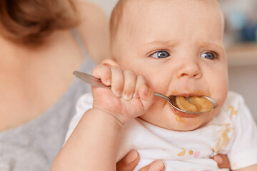 Close up portrait of cute toddler girl trying to eat fruit or vegetable puree with spoon, unknown woman holding feeding infant baby, feeding up, indoor shot.