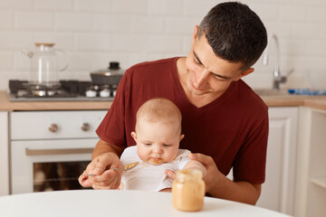 Obraz na płótnie Canvas Indoor shot of handsome father wearing burgundy t shirt with charming infant daughter. man feeding his baby with puree, posing in light room against kitchen set.