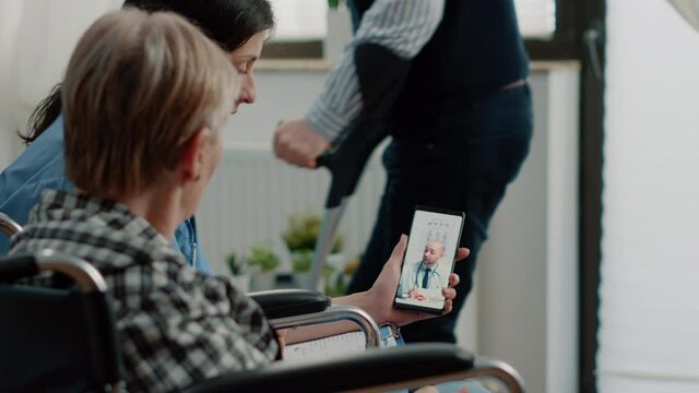 Senior invalid patient talking to doctor on video call in nursing home. Old woman with disability in wheelchair using telemedicine and online conference while nurse holding smartphone