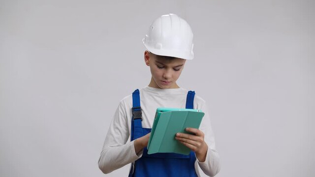 Focused serious boy in hard hat and overalls using tablet thinking and looking at camera. Portrait of confident thoughtful Caucasian child in builder uniform posing at white background. Foreman job