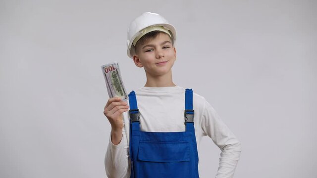 Boy in builder contractor uniform counting money looking at camera with satisfied facial expression smiling. Portrait of proud Caucasian kid choosing profitable profession posing at white background