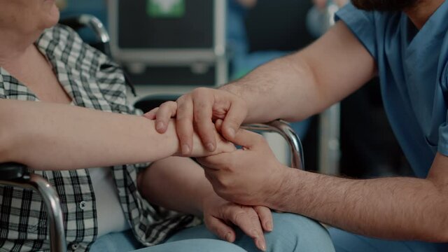 Close up of hands of invalid senior woman and man nurse in nursing home. Medical assistant giving support and comforting retired person, caregiver helping with therapy and recovery