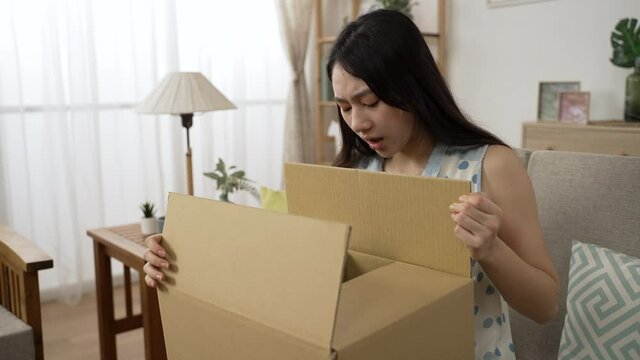 disappointed asian woman carrying and sitting on couch at home to unpack package box is looking at the damaged item feeling unhappy with the bad delivery service
