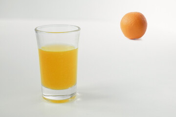 A glass of orange juice in the foreground and a defocused orange in the background.