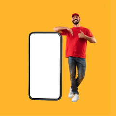 Middle-Eastern Guy Standing Near Big Smartphone Gesturing Thumbs-Up, Yellow Background