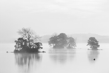 Three spooky black white islands on calm, misty lake water