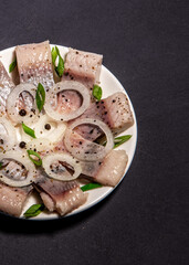 Fillet of salted herring with spices and onions on a plate. Salted sliced ​​fish on a dark background. Foods with healthy unsaturated fats, omega-3s. Top view with copy space. Healthy food