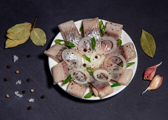 Fototapeta na wymiar Fillet of salted herring with spices and onions on a plate. Salted sliced ​​fish on a dark background. Foods with healthy unsaturated fats, omega-3s. Top view with copy space. Healthy food