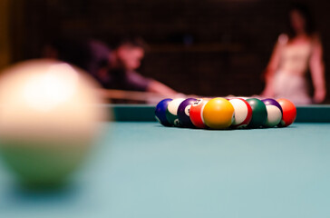 cue ball in front of all other billiard balls on green billiard table. a couple in love playing pool in the background out of focus.