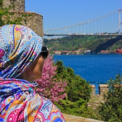 Focus on foreground.Elegant mature caucasian woman traveler in traditional bright oriental headscarf looking to Fatih Sultan Mehmet Bridge on Bosphorus in famous Rumeli Hisari Fortress,Istanbul,Turkey