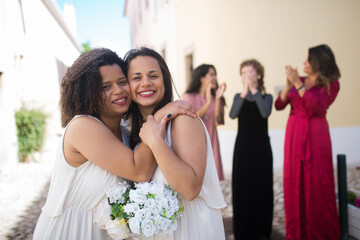 Portrait of cute smiling brides. Two young women hugging, looking at camera. Female guests laughing and applauding in background. Wedding, LGBT, celebration concept
