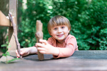 Little preschool boy sitting near a wooden bank in a forest and playing with sticks. Child spending time outdoor in summer. Toddler in a park. Ecological playground.