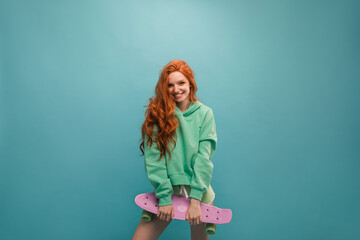 Young red-haired girl with embarrassed look smiles at camera on blue background. She has long wavy hair and light-colored clothing. In hands of fashionable lilac skateboard. Concept active recreation.