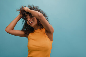 Young african brunette holding her curly hair and feeling pretty good against blue background. Her eyes are closed, wearing bright yellow top. Positive emotion concept