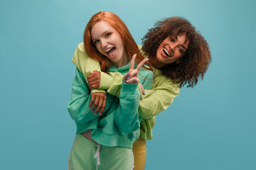Dark-skinned girl hugs her fair-skinned friend from behind on blue background in studio. They have smile with open mouth. Redhead shows peace with her left hand. - obrazy, fototapety, plakaty