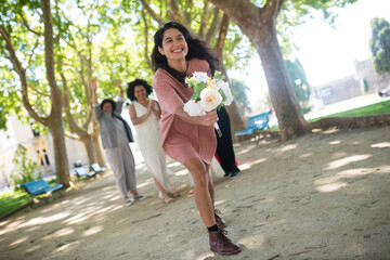 Fototapeta na wymiar Smiling young woman catching wedding bouquet. Dark haired woman in pink dress with flowers. Women of different nationalities in festive dresses laughing in background. LGBT wedding celebration concept
