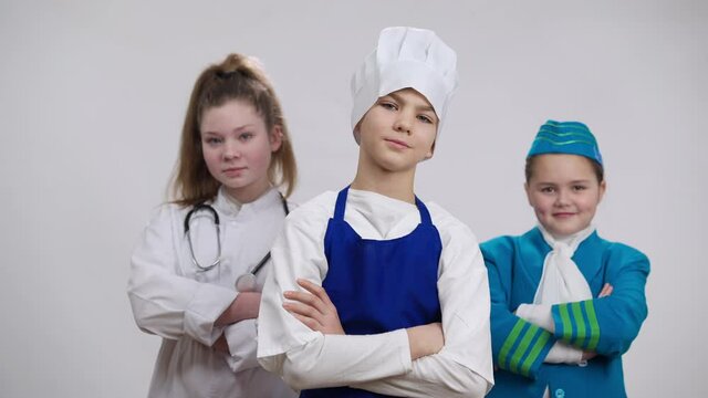 Portrait Of Confident Boy In Chef Cook Uniform Posing Crossing Hands Looking At Camera With Girls In Stewardess And Doctor Outfit At Background. Children Choosing Professions At Background