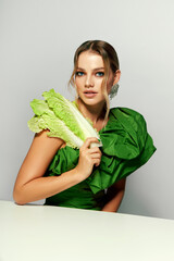 Portrait of a beautiful young woman with bright makeup and in a bright green dress. Woman holding cabbage leaves. The concept of ecological products, cleansing, detoxification. Fashionable green dress