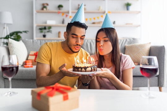 Positive interracial couple in party hats blowing lit candles on birthday cake, drinking wine, exchanging gifts at home - Powered by Adobe