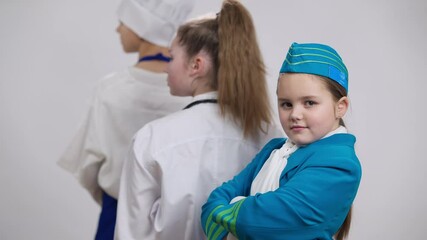 Cute girl flight attendant turning crossing hands looking at camera with little doctor and chef cook at background. Portrait of pretty Caucasian child choosing aviation occupation posing