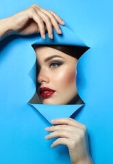 The face of a young beautiful woman with bright makeup and plump red lips looks through the blue bmagi. Advertising of decorative and care cosmetics. Advertising concept.
