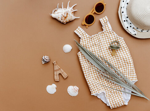 Summer kids accessories on brown background. Cute retro swimsuit, straw hat, sunglasses and seashells, summer concept flat lay