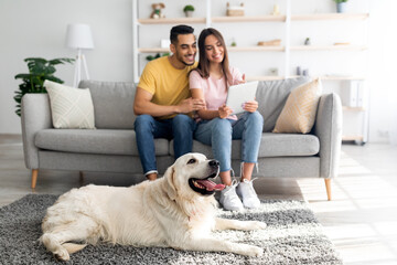 Affectionate diverse couple using tablet on couch, cute pet dog lying on the floor at home, selective focus