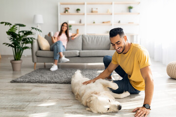 Happy Arab guy stroking cute dog on floor at home, his girlfriend relaxing with cup of coffee on background, copy space