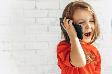 Cute little girl talking on a cell phone in a red T-shirt on a white background. Digital technologies in the hands of a child. Communication at a distance.