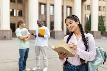 Happy university student girl holding book, standing outdoors at university campus with her classmates on background