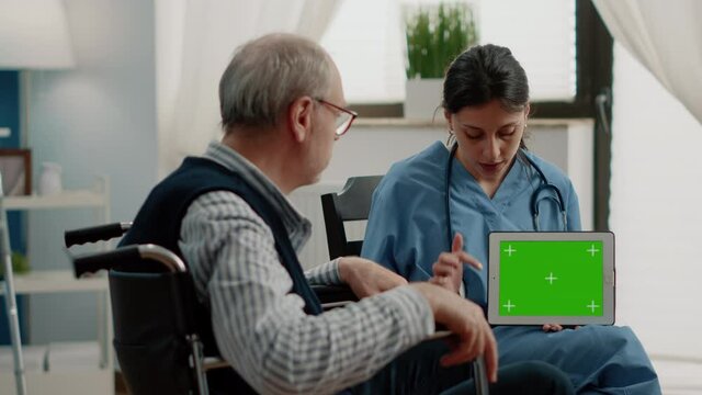 Medical assistant holding tablet with horizontal green screen while talking to invalid patient. Nurse showing chroma key with mockup template and isolated background to old man in wheelchair