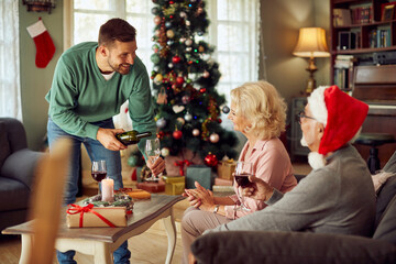 Happy man serves wine to senior parents while celebrating Christmas together at home.