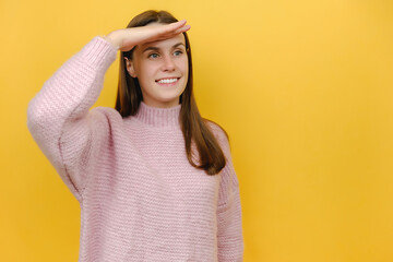 Portrait of smiling optimistic waiting dreamful happy cute young woman 20s holding hand at forehead looking far away distance, dressed in pink knitted sweater, isolated on yellow studio background