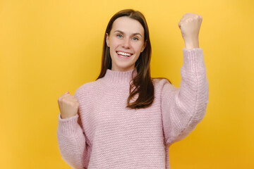 Happy excited successful overjoyed lucky attractive pretty young female 20s do winner gesture clench fists celebrating, dressed in knitted sweater, isolated on yellow color background wall in studio