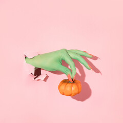 Halloween creative layout with green witch hand with bright pink nails holding pumpkin against pastel pink background. Aesthetic season idea. Minimal Halloween concept.