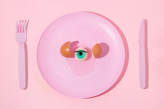 Halloween creative layout with pastel pink plate, fork,knife, eyeball and eggshell on pastel pink background. Aesthetic fashion food restaurant concept. Minimal surreal breakfast idea.