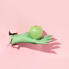 Halloween creative layout with green witch hand with bright pink nails holding apple against pastel...