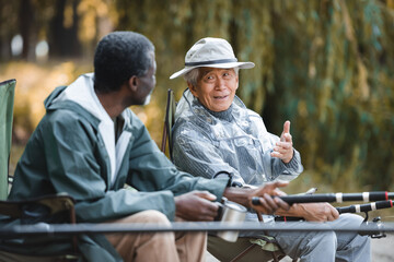 Naklejka na ściany i meble Senior asian man talking to african american friend while fishing outdoors