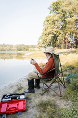 Senior man in fishing outfit holding thermo cup near toolbox and lake