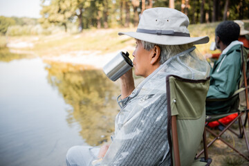 Senior asian man holding thermo cup near blurred friends in fishing outfit near lake