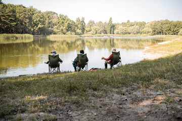 Obraz na płótnie Canvas Interracial senior men in fishing outfit sitting near lake in park