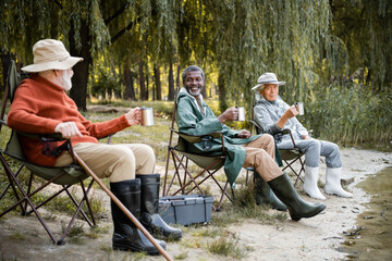 Smiling multicultural senior men in fishing outfit with thermo cups sitting near lake in park