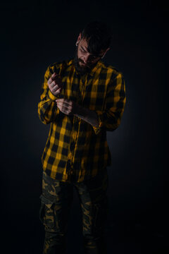 Dark image of a bearded man with tattoos fixing his buttons on his yellow and black checkered shirt