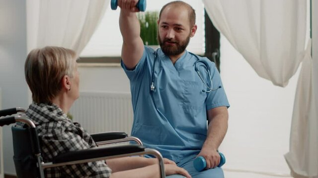 Medical assistant giving assistance to disabled old woman for physical exercises with dumbbells in nursing home. Nurse helping senior patient in wheelchair with activity for recovery