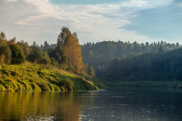 Fototapeta premium Bend of the river. The banks are covered with green grass and forest. The autumn trees began to turn yellow. Fog in the distance. Blue sky with some clouds. Sunny day. Nikola-Lenivets park. Ugra river