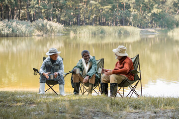 Positive interracial men in fishing outfit holding thermo cups on chairs on lake coast