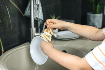 A child washes dishes with a wooden brush with natural bristles in the kitchen.