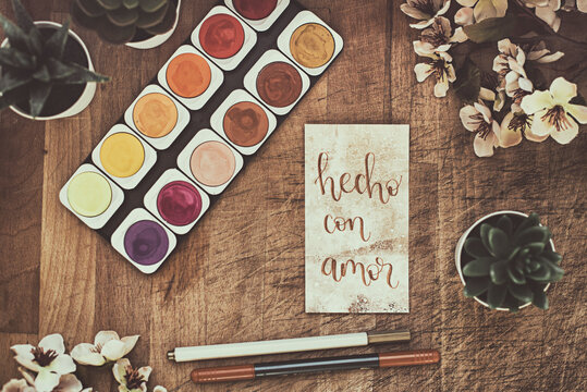 Watercolour paints on a table with pot plants, flowers and a card with the words hecho con amor